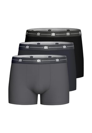 Lihaksikas mies pukeutuneena tummanharmaisiin bokserishortseihin seisoo kädet lanteilla yksiväristä vaaleaa taustaa vasten.