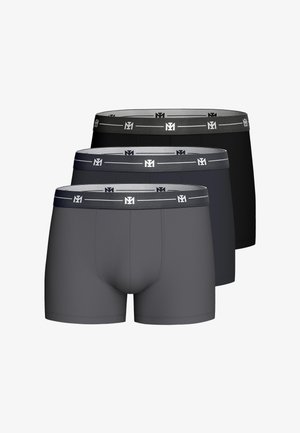 Gespierde man die donkere grijze boxershort draagt, staand met handen in de zij tegen een effen lichte achtergrond.