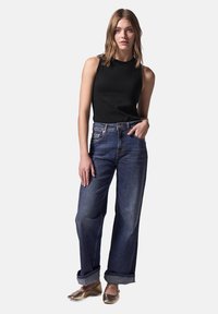 Top negro sin mangas combinado con vaqueros de pierna ancha de talle alto en azul oscuro, remangados en el dobladillo, y zapatos planos metálicos dorados. Textura suave y ajuste casual.
