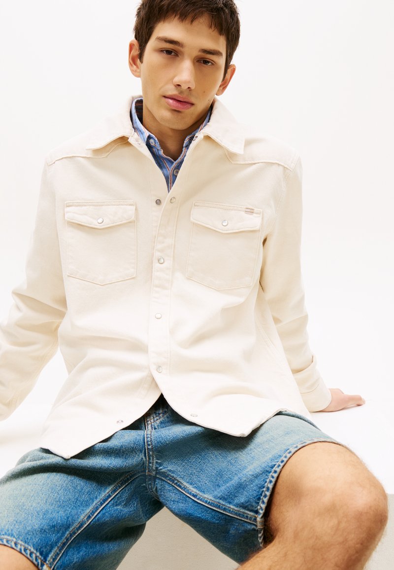 Jonge man zittend, draagt een beige overhemd met knopen over een blauw gestreept shirt en spijkershorts, tegen een effen witte achtergrond.