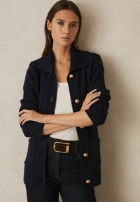 Cardigan de tricot bleumarin cu guler rib și nasturi mari aurii, două buzunare frontale și mâneci texturate, purtat cu un top alb și blugi negri.