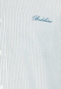 Gros plan sur la poche d'une chemise blanche à rayures bleu clair avec le texte brodé en bleu « Backslime » au-dessus de la poche.