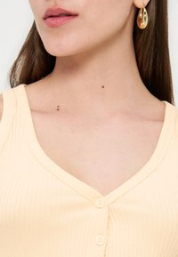 Κοντινή λήψη μιας γυναίκας που φοράει ανοιχτό κίτρινο ριμπ αμάνικο τοπ με κουμπιά και χρυσό στριφτό κρίκο στο αυτί.