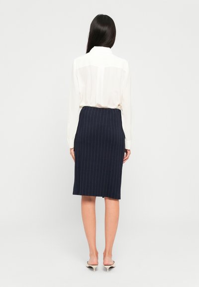 Vrouw met lang, donker haar, gekleed in een witte blouse met lange mouwen, een marineblauwe pinstripe rok tot de knieën en witte, hoge muilen, staande met haar rug naar de kijker.