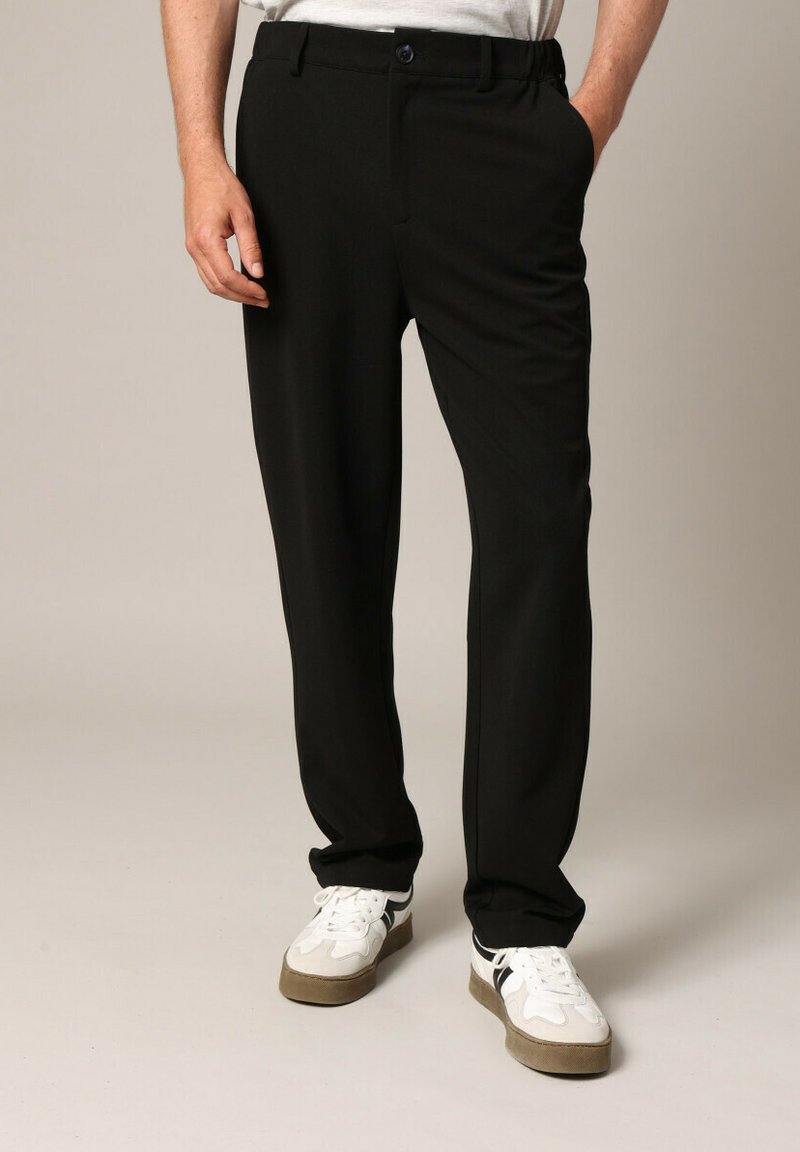 Zwarte broek van stof met een gladde textuur, voorzien van een knoop- en ritssluiting, zijzakken en een aansluitende pasvorm, gecombineerd met witte sneakers.