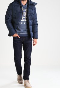 Granatowa kurtka puchowa z zamkiem z przodu, pikowaną fakturą i kapturem, noszona na niebieskiej bluzie i ciemnych dżinsach, w połączeniu z jasnobrązowymi butami.