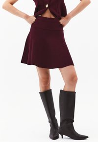 Burgundy neulehame, jossa on leveä helma ja leveä vyötärö, yhdistettynä polveen saakka ulottuviin mustiin nahkasaappaisiin, joissa on terävä kärki.