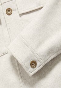 Mankiet jasnobeżowej wełnianej kurtki o gładkiej fakturze z pojedynczym brązowym guzikiem na cztery dziurki do zapinania.