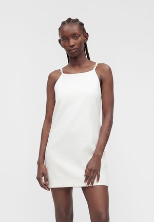 Jeune femme aux cheveux tressés portant une mini-robe blanche sans manches avec de fines bretelles, se tenant devant un fond clair uni.