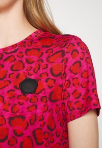 Chemise rose à manches courtes avec un motif léopard rouge et noir. Comprend un écusson rond noir avec logo sur le côté gauche de la poitrine.