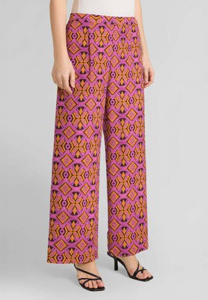 Donna che indossa pantaloni a gamba larga con motivo floreale geometrico rosa, arancione e nero e sandali neri con tacco e cinturini.