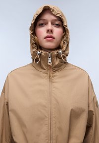 Veste légère marron avec un col haut et une capuche réglable. Présente une fermeture zippée et des accents à cordon à la capuche. Texture lisse.
