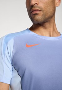 Mann iført lyseblå Nike sportstrøye med oransje swoosh-logo, kortet ved nakke og skulder mot ensfarget bakgrunn.