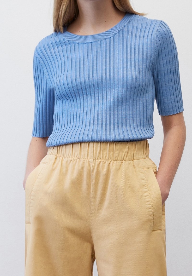 Světle modrý žebrovaný top s krátkým rukávem a kulatým výstřihem, spárovaný s vysokými žlutými kalhotami, které mají boční kapsy a strukturovanou látku.