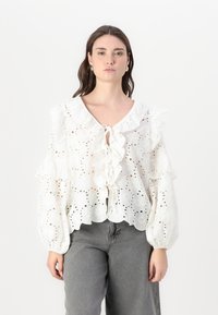 White cotton blouse with eyelet pattern, ruffled neckline and sleeves, tie closure, and scalloped hem. Paired with gray wide-leg jeans.
