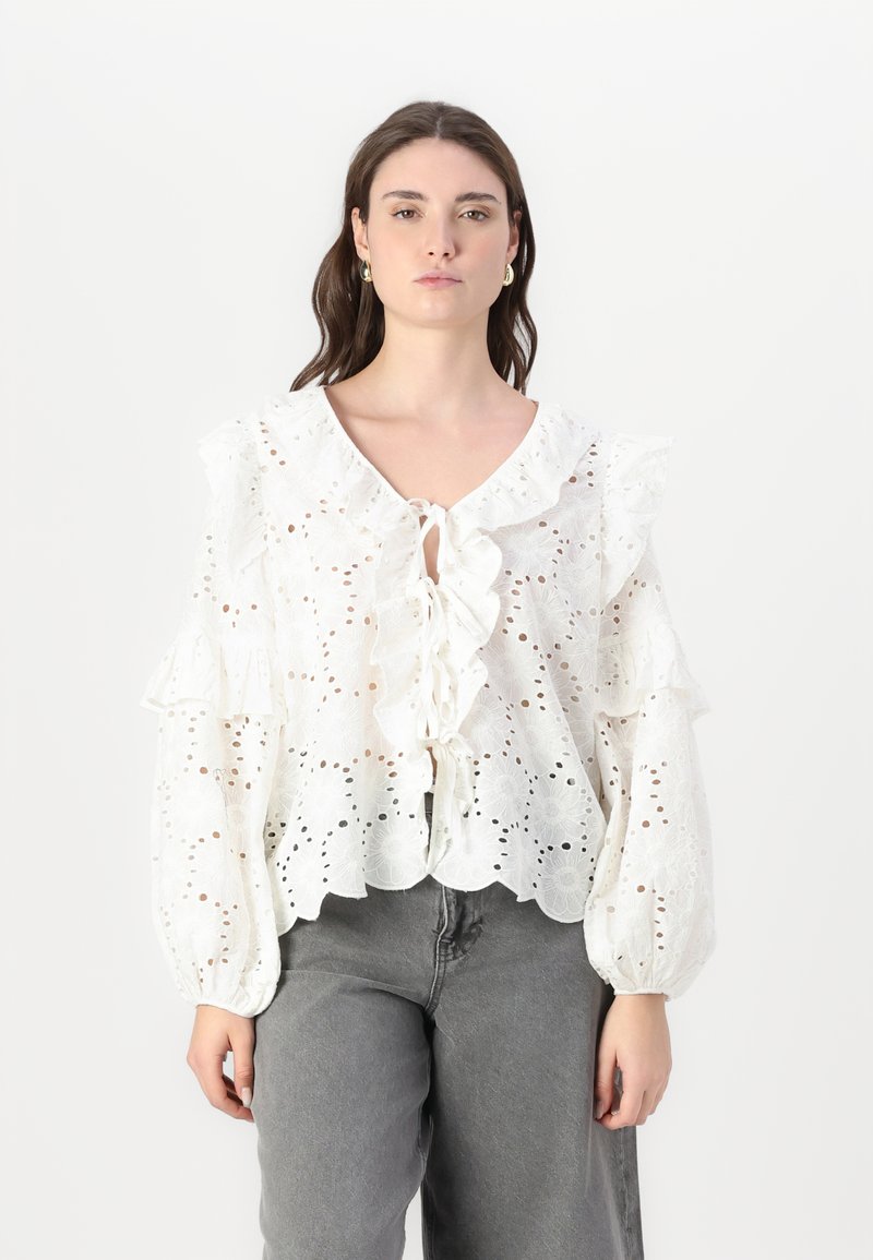 White cotton blouse with eyelet pattern, ruffled neckline and sleeves, tie closure, and scalloped hem. Paired with gray wide-leg jeans.