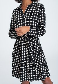 Robe chemise noire et crème à pois, avec un col, un boutonnage sur le devant, des manches longues et un détail de taille nouée. Texture de tissu lisse.