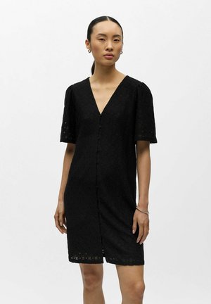 Kvinne med rett svart hår som har på seg en kortermet, knelang svart kjole med knappestolpe og subtilt teksturmønster, samt ørepynt med ringer.