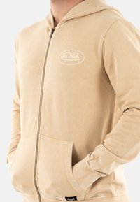 Felpa khaki con zip realizzata in morbido misto cotone, dotata di logo frontale, tasche a marsupio e polsini a costine. Texture liscia con una vestibilità rilassata.