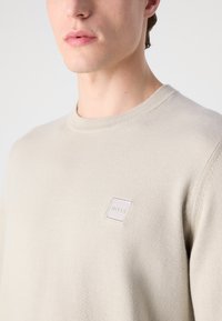 Bež pleten pulover s krojnim ovratnikom, ki ima kvadratno logotipno obliž v svetlejši odtenek na levem prsnem predelu. Gladka tekstura, lahkoten material.