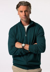 Tyrkysový zipový svetr s žebrovanými manžetami a lemem, vyrobený z měkké tkaniny. Na zápěstí je nasazen stříbrný hodinky s hnědým koženým páskem.