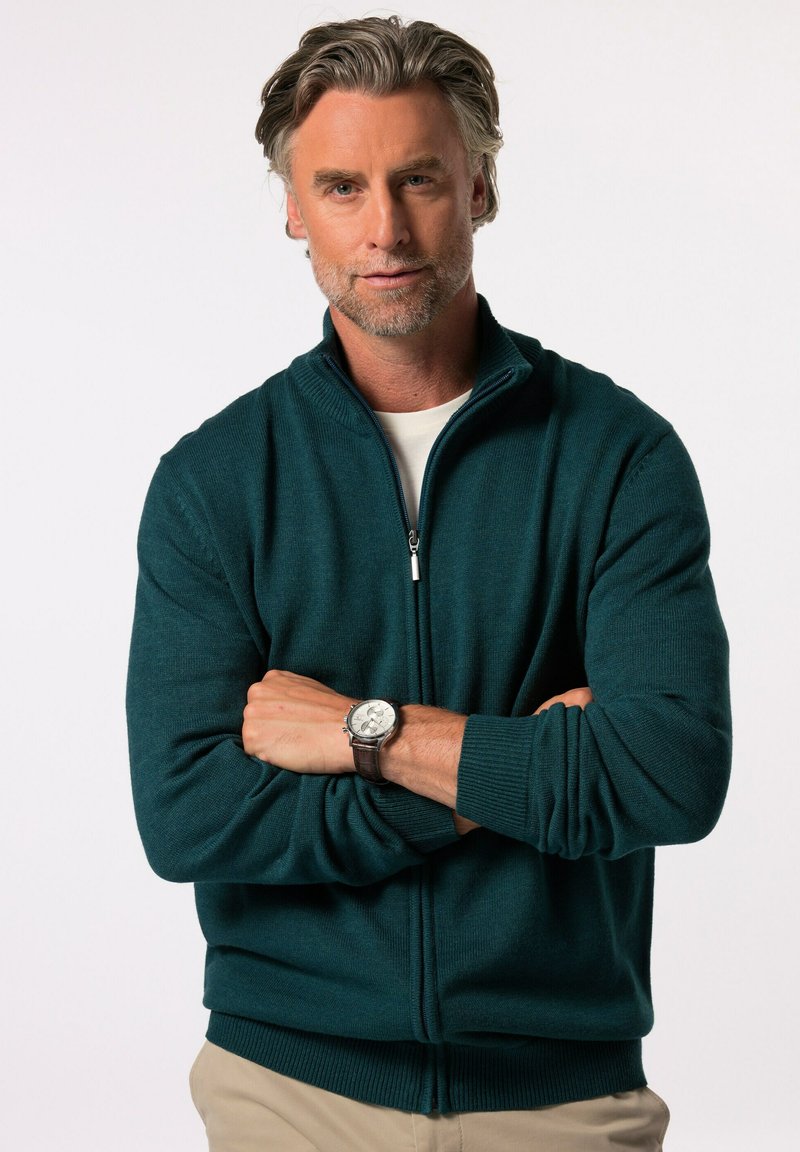 Tyrkysový zipový svetr s žebrovanými manžetami a lemem, vyrobený z měkké tkaniny. Na zápěstí je nasazen stříbrný hodinky s hnědým koženým páskem.