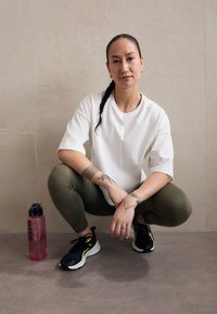 Športový outfit pozostávajúci z bieleho trička, olivovo zelených legín a čiernych tenisiek s neónovými akcentmi; vedľa leží ružová fľaša na vodu.