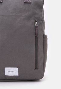 Sac à dos en tissu gris présentant une poche zippée, une forme rectangulaire et une étiquette avec le nom de la marque sur un fond blanc.