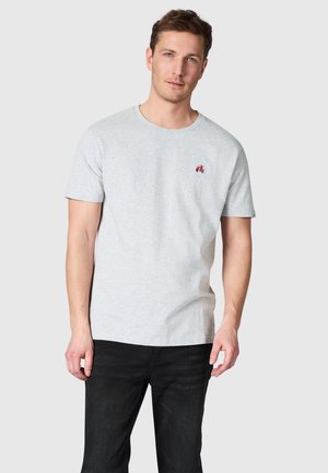 Man draagt een lichtgrijs t-shirt met korte mouwen met een klein rood motorfietsembleem op de linkerborst en zwarte jeans, staand tegen een effen achtergrond.