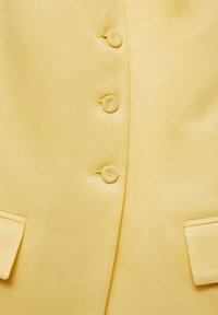 Gros plan d'un blazer jaune avec trois boutons sur le devant et deux poches à rabat près du bas.