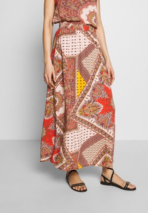 Femme portant une jupe longue et fluide avec des motifs cachemire et géométriques rouges, jaunes et beiges, associée à des sandales noires à lanières.