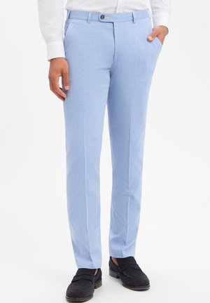 Hellblaue, maßgeschneiderte Hose, kombiniert mit schwarzen Loafers und einem weißen Hemd, eine Hand in der Tasche, die andere locker am Bein.