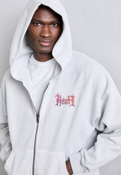 Grå zip-up hoodie med en framficka, en mjuk textur och en flerfärgad flammig grafisk logotyp på bröstet. Har en bred huva.
