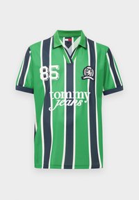 Green polo shirt with white and navy vertical stripes, "85" number, "tommy jeans" text, and a navy embroidered crest on the chest.