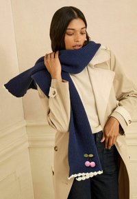 Strikket marineblå tørklæde med hvide frynser, der har lyserøde pom-pom kirsebær og grønne blade som dekorative detaljer, holdt af en model.