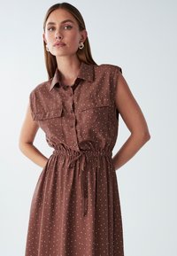 Brązowa sukienka z guzikiem, krótki rękaw, białe kropki, dwie kieszenie na piersi oraz elastyczny pas z detalem wiązania. Gładka tekstura materiału.