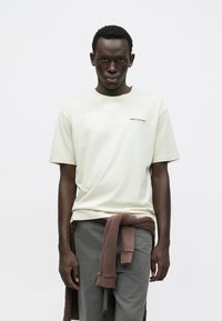 Gaiši bēša kokvilnas T-krekls ar īsām piedurknēm un McLaren logotipu; kopā ar pelēkām biksēm un brūnu džemperi, kas sasiets ap vidukli.