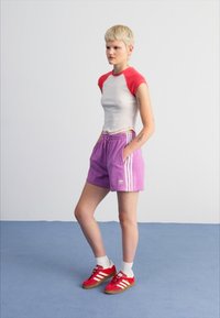 Rød og hvit topp med korte ermer, lilla shorts med hvite striper, og røde joggesko med hvite detaljer. Glatt stoff, avslappet passform.