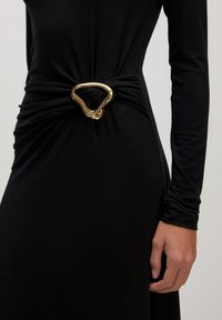 Zwart langemouwen jurk met geplooide stof bij de taille, voorzien van een unieke, onregelmatige gouden metalen gesp als accent. Over het geheel een gladde textuur.