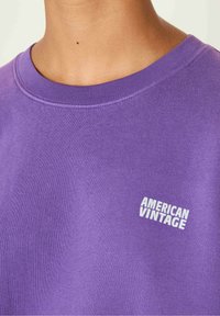 Violetti huppari, joka on valmistettu pehmeästä kankaasta, ja jossa on pyöreä kaula-aukko sekä valkoinen "AMERICAN VINTAGE" -logo vasemmassa rinnassa.