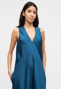 Γυναίκα με μακριά σκούρα μαλλιά φορώντας αμάνικο τιρκουάζ σατέν φόρεμα με βαθύ V λαιμόκοψη, με τα χέρια στις πλαϊνές τσέπες, κοιτάζοντας την κάμερα μπροστά σε απλό φόντο.