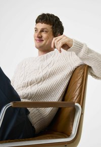 Mann mit kurzen braunen Haaren, der einen cremefarbenen Pullover mit Zopfmuster trägt und entspannt in einem braunen Ledersessel mit Metallarmlehnen sitzt.