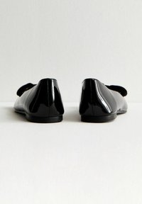 Zapatos planos de charol negro brillante vistos desde la parte posterior, colocados sobre una superficie blanca con un fondo sencillo.