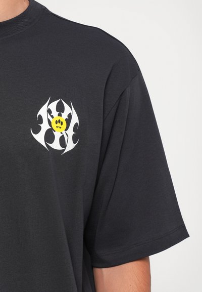 Camiseta negra de manga corta con un pequeño diseño abstracto en blanco y amarillo en el lado izquierdo del pecho que presenta un círculo amarillo con tres ojos negros y colmillos.