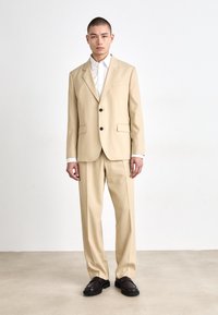 Beige skræddersyet jakkesæt med en enkeltstående jakke, notch-lapel og knaplukning. Parret med en hvid skjorte og brune sko.