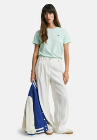 Muntgroene t-shirt, losse witte gestreepte broek, houdt een blauwe varsityjack met witte accenten vast. Casual stijl, zachte texturen.
