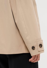 Beež trench coat'i varrukas, mis on varustatud nööpidega mansetiga, kus on kaks tumedat nööpi. Kangas näeb välja siledana ja veidi tekstureerituna.