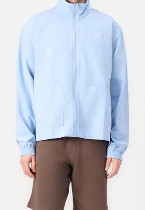 Giacca azzurro chiaro con cerniera, maniche lunghe e due tasche frontali indossata con pantaloncini marroni su una persona in piedi contro uno sfondo bianco.