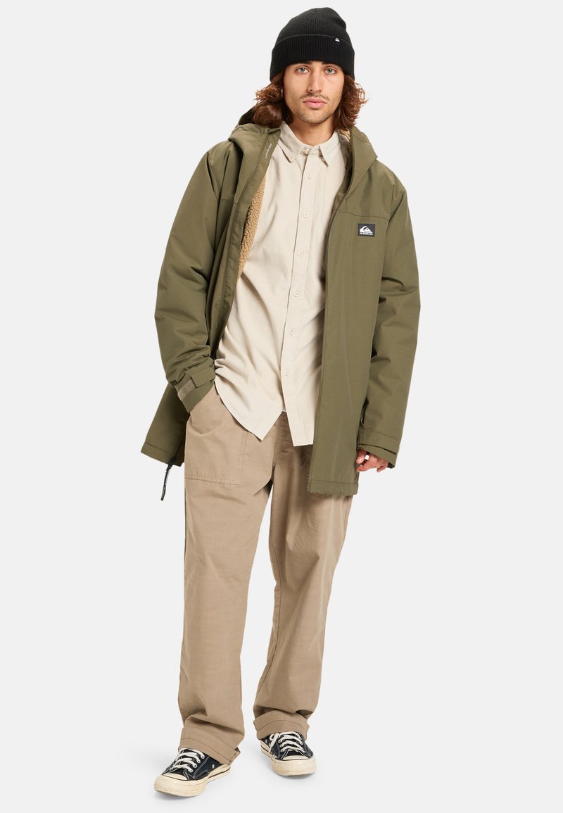 Groene geïsoleerde jas met een bontvoering capuchon, beige overhemd met knopen, tan cargobroek en zwarte canvas sneakers.