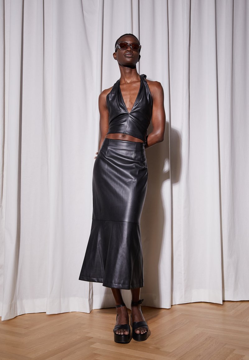Top halter de couro preto e saia midi com corte evasê, textura suave, design ajustado, com sandálias plataforma a condizer, contra um fundo claro.